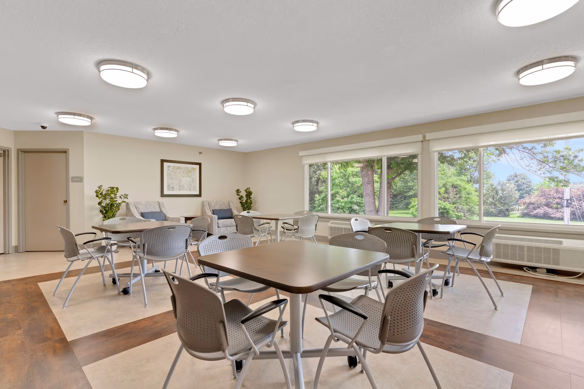 A bright and spacious common area with several square tables and gray chairs arranged around them. Large windows on one side offer a view of green trees and a lawn outside. The room has light-colored walls, ceiling lights, and a couple of armchairs with cushions near the back wall, along with some potted plants and a framed picture.