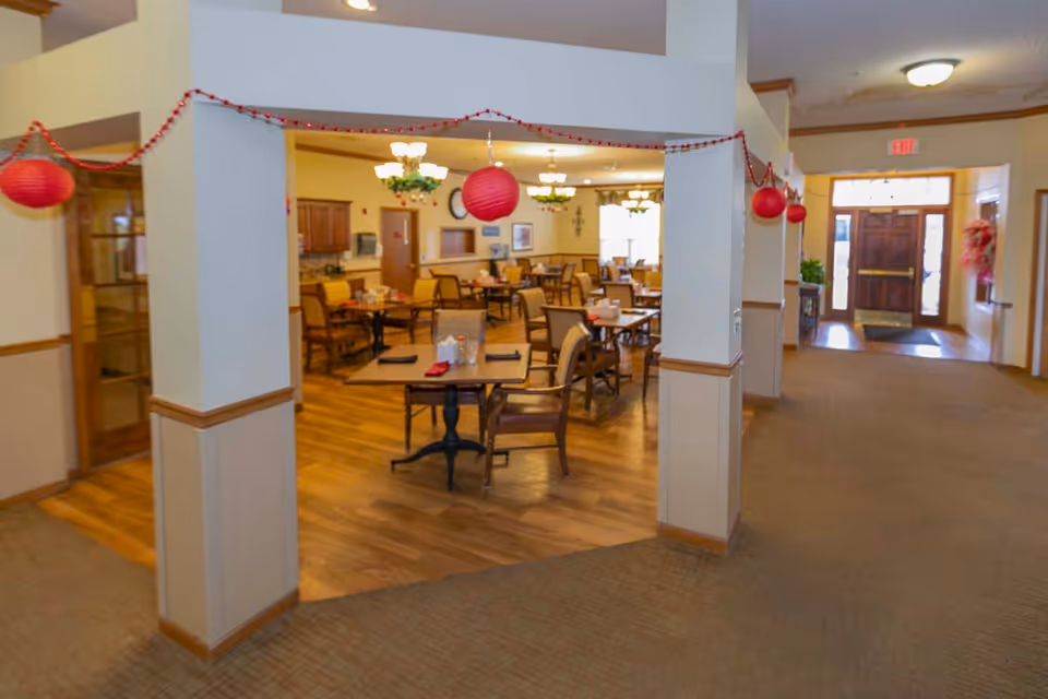 Interior view of a senior living facility dining room with wooden floors, multiple tables and chairs arranged for dining, red hanging lantern decorations, and warm lighting fixtures. The entrance door is visible in the background along a carpeted hallway.