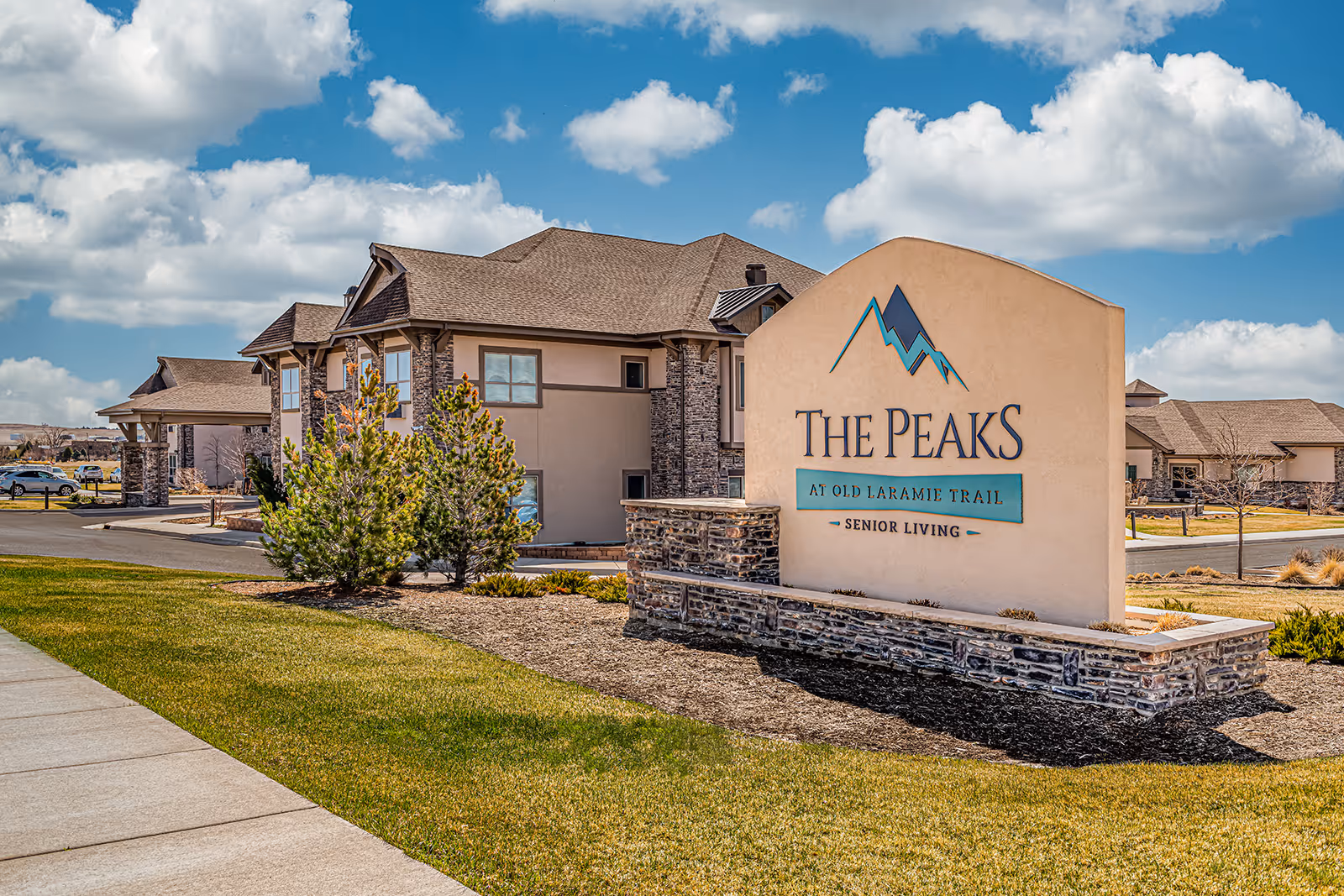 Exterior view of The Peaks at Old Laramie Trail senior living facility with a large sign in the foreground and buildings with stone and beige siding in the background under a partly cloudy blue sky.