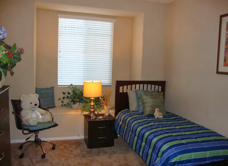 A small bedroom with a single bed covered in a blue and green striped bedspread, a small stuffed animal on the bed, a nightstand with a lamp and decorative items, a window with closed blinds, a chair with a teddy bear on it, and a framed picture on the wall.