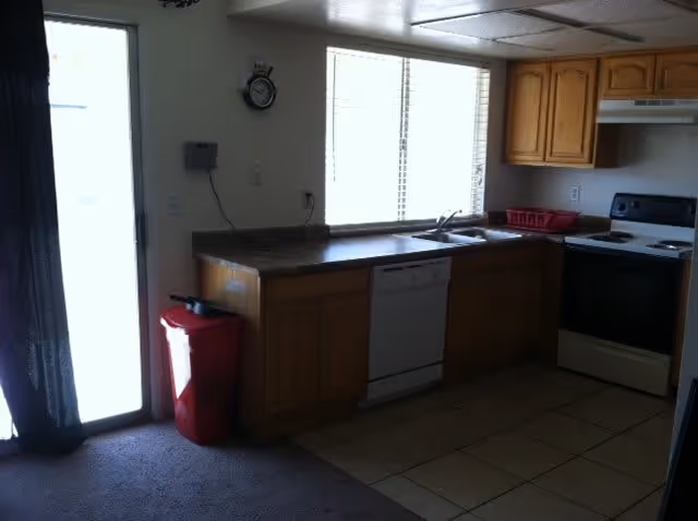 A kitchen area with wooden cabinets, a countertop, a white dishwasher, a stove with an oven, a red dish rack, a red trash bin, and a window with blinds letting in natural light.
