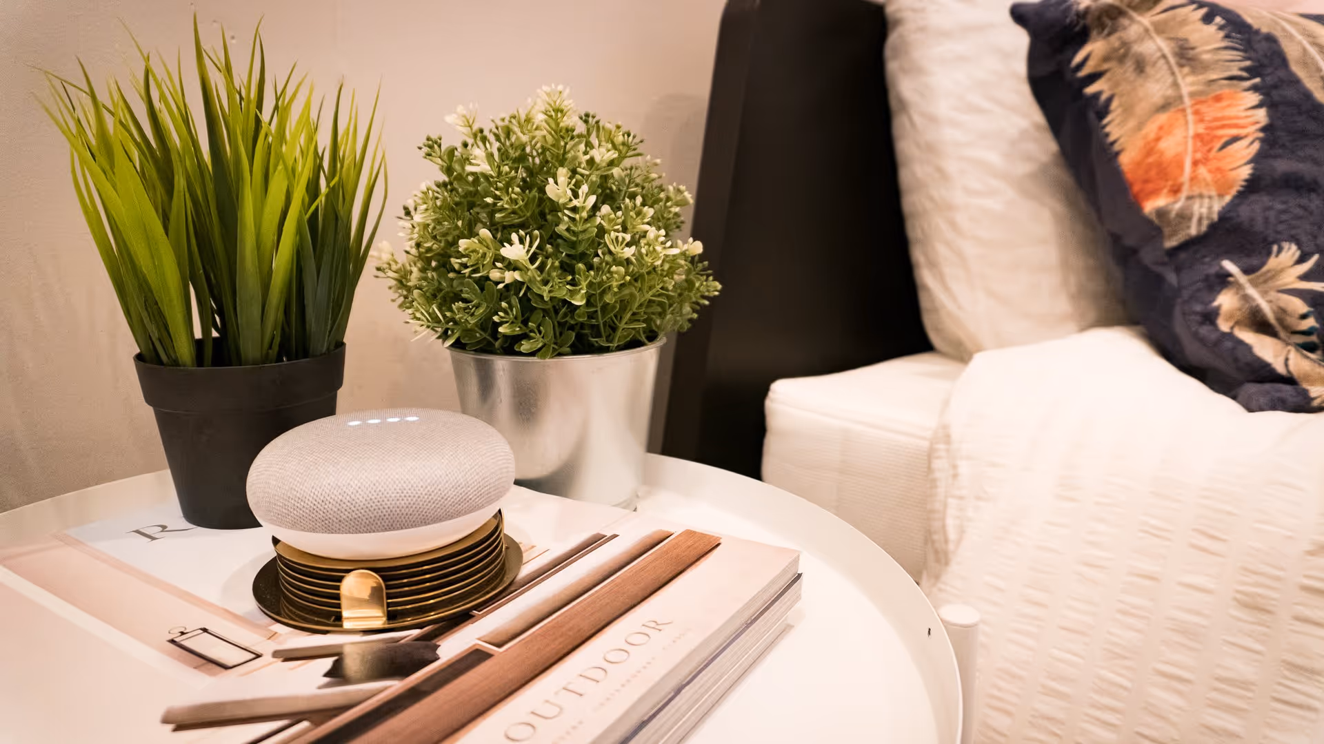 Nightstand holding two potted plants, a smart speaker, and a book beside a bed with pillows.