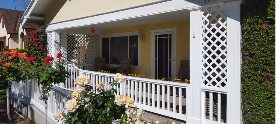 A bright and inviting porch area of a residential building with white railings and lattice panels. The porch is decorated with potted flowers along the railing and has comfortable seating with cushioned chairs. Surrounding the porch are blooming rose bushes with red, orange, and cream-colored flowers, and a green hedge on the right side.