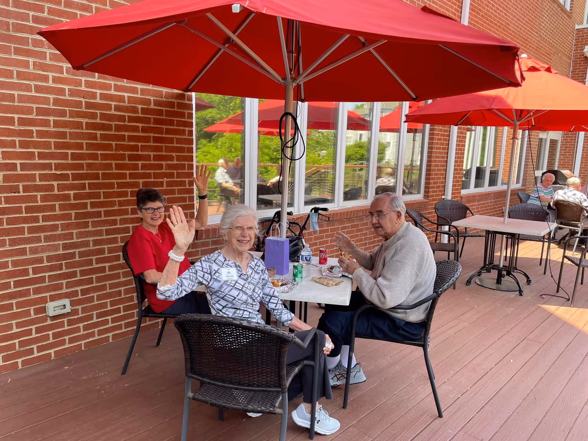 Three elderly people sitting at a table under a red umbrella on a wooden patio outside a brick building. Two women and one man are smiling and waving at the camera. There are other tables and chairs with red umbrellas in the background, and large windows reflect the outdoor scene.