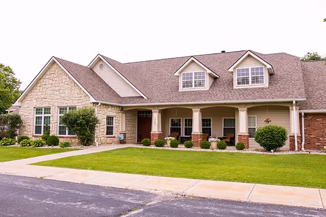 Exterior view of a single-story residential building with a stone and brick facade, a covered porch with columns, multiple windows, and a well-maintained lawn and shrubs in front.