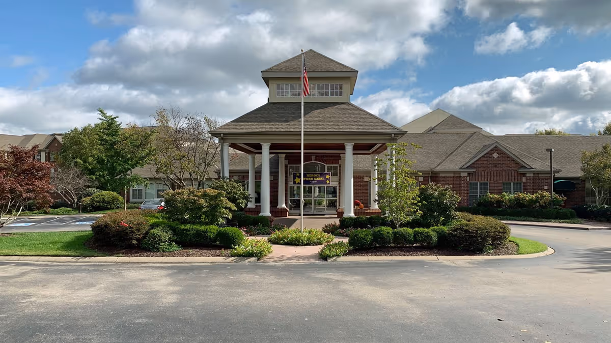 Front exterior view of a senior living facility building with a covered entrance supported by white columns, an American flag on a flagpole in front, surrounded by well-maintained bushes and trees under a partly cloudy sky.
