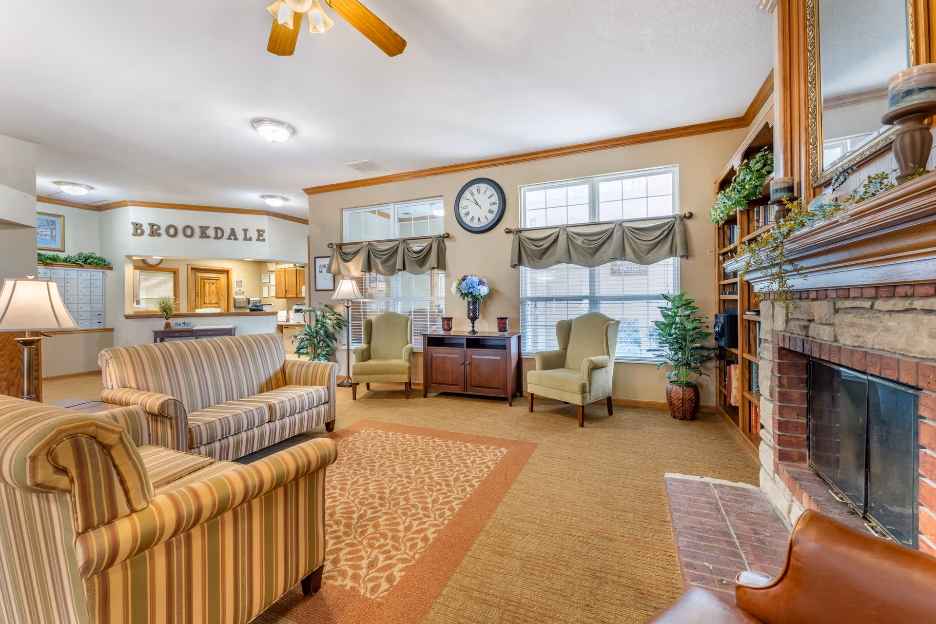 A cozy living room area in Brookdale Hays featuring striped sofas, two green armchairs, a wooden cabinet with a vase of flowers, a large wall clock, and a stone fireplace with a wooden mantel decorated with plants and candles. The reception area with the word 'BROOKDALE' on the wall is visible in the background.