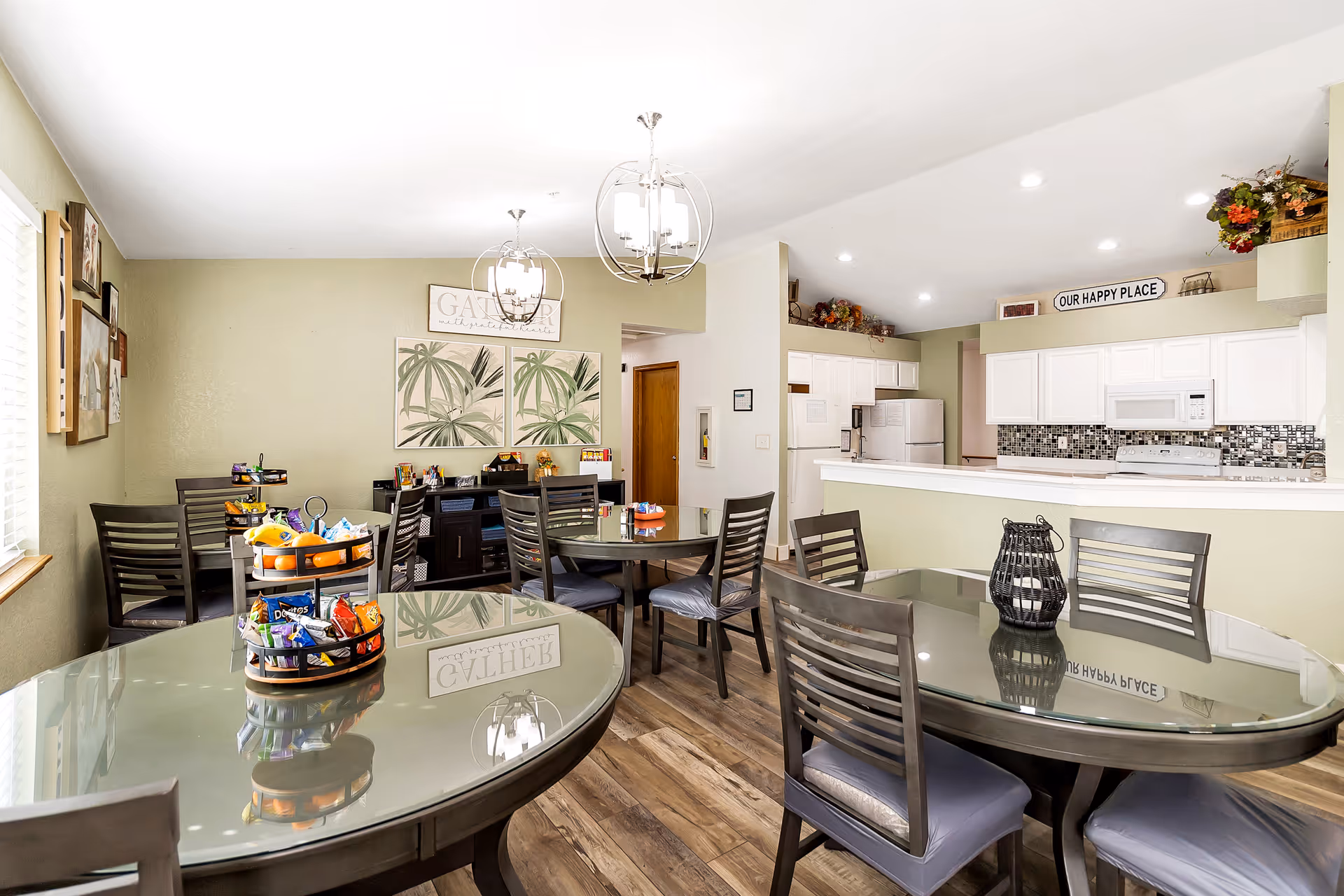 A bright dining area in Ashley Manor Memory Care featuring multiple round glass-top tables with dark wooden chairs. The room has light green walls decorated with framed botanical prints and a sign that reads 'GATHER'. The kitchen area is visible in the background with white cabinets, a mosaic tile backsplash, and a sign above that says 'OUR HAPPY PLACE'. Snacks are arranged on a tiered tray on one of the tables. The floor is wood-style laminate, and modern pendant lights hang from the ceiling.