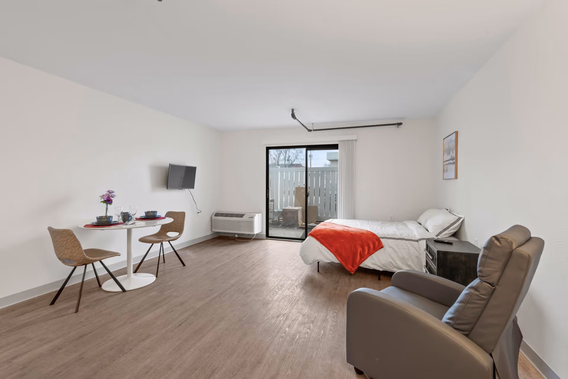 A bright, spacious room with a bed covered in white bedding and a red throw blanket, a small round dining table set for two with chairs, a wall-mounted TV, a comfortable armchair, and a sliding glass door leading to an outdoor patio area.