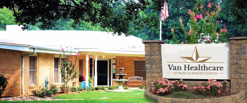 Exterior view of Van Healthcare nursing and rehabilitation facility showing a single-story brick building with a covered entrance, a water fountain, a bench, and landscaped greenery including trees and flowering plants. A stone sign with the facility name is prominently displayed in the foreground.