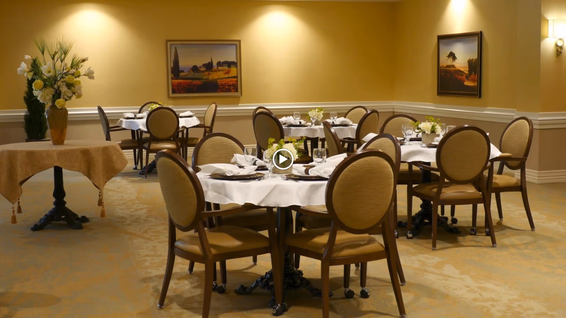 A dining room with several round tables covered with white tablecloths, each set with plates, glasses, and napkins. The chairs around the tables have cushioned backs and seats. The room has beige walls adorned with framed landscape paintings and a table with a large floral arrangement in a vase.