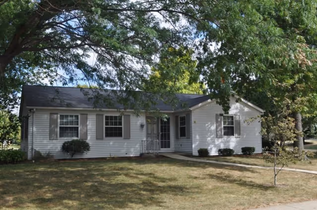 Single-story white house with gray shutters surrounded by trees and a grassy yard under a partly cloudy sky.
