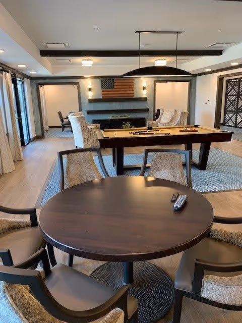 A spacious common area featuring a round wooden table with four cushioned chairs in the foreground, a pool table in the middle, and a seating area with armchairs near a wall-mounted American flag and a modern fireplace. The room has large windows with curtains, wooden flooring, and contemporary lighting fixtures.