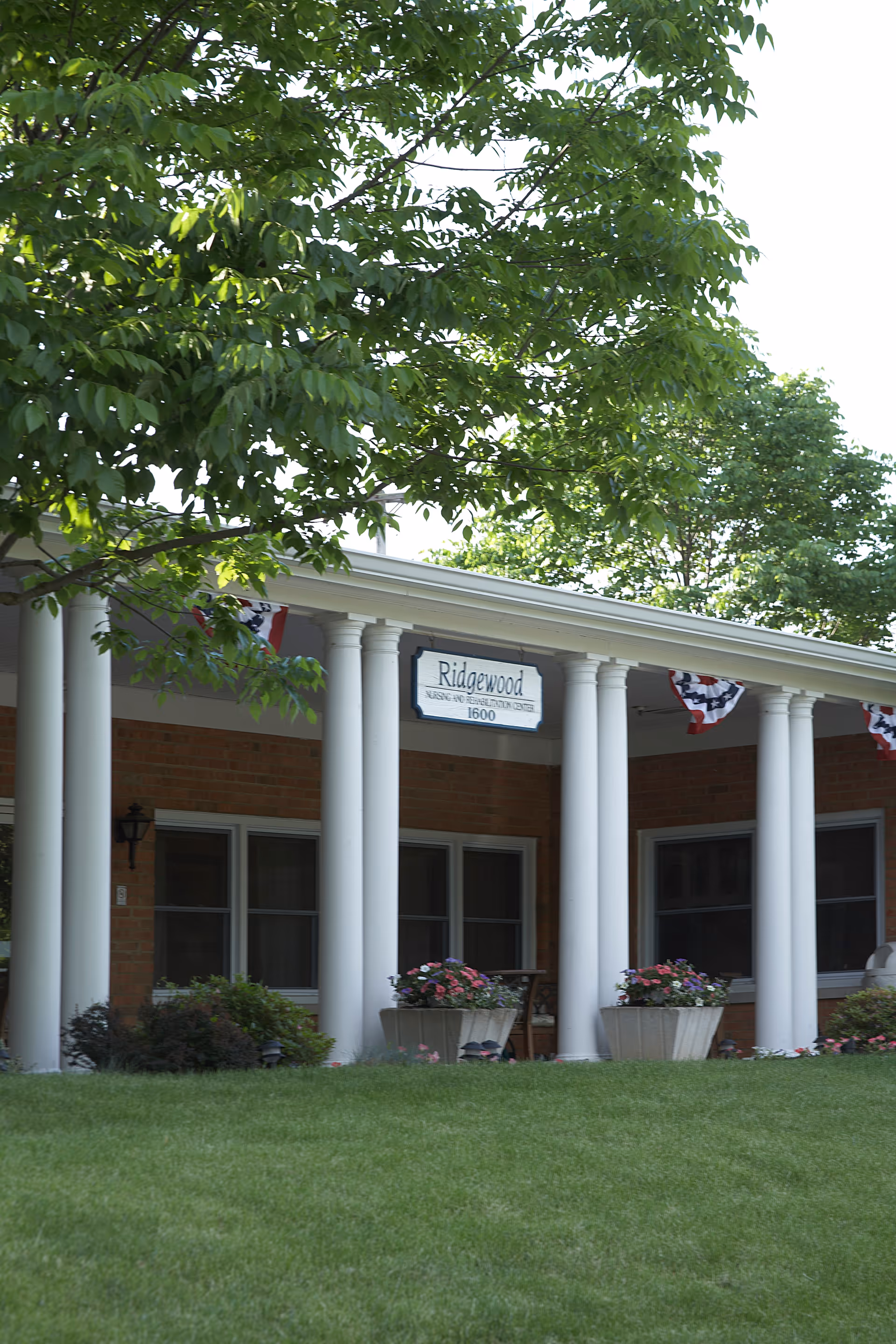 Front entrance of a brick nursing facility with white columns, a 'Ridgewood' sign, potted flowers, and a green lawn.
