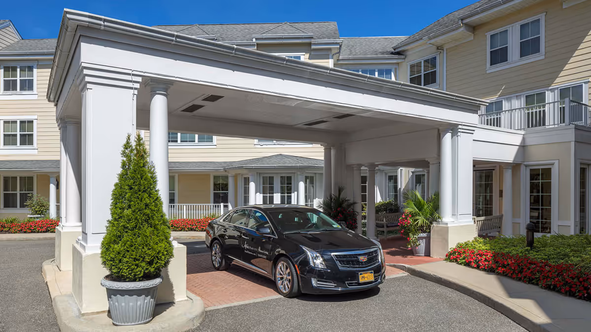 Entrance of Atria Plainview senior living facility with a covered drop-off area supported by white columns, a black Cadillac car parked under the canopy, potted plants, and well-maintained flower beds surrounding the building.