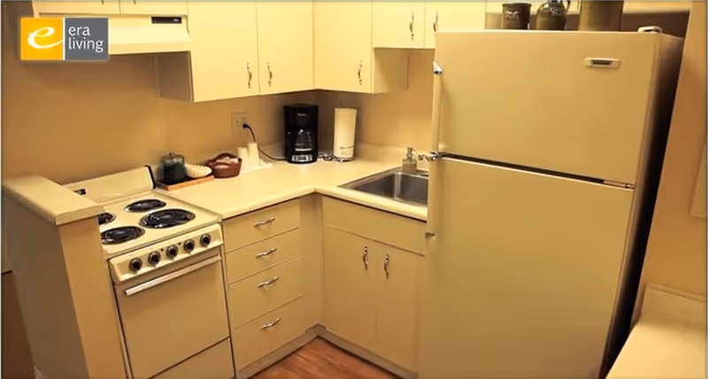 A compact kitchen with cream-colored cabinets and appliances including an electric stove, refrigerator, and a stainless steel sink. There is a coffee maker and some kitchen items on the countertop. The floor is wooden.