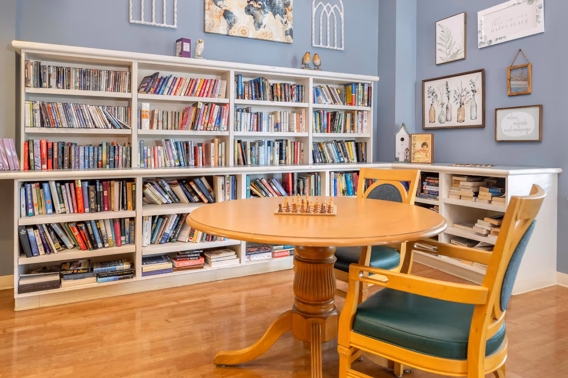 A cozy reading and game area with a round wooden table and two wooden chairs with green cushions. Behind the table is a large white bookshelf filled with books. The walls are painted blue and decorated with framed artwork and decorative items.