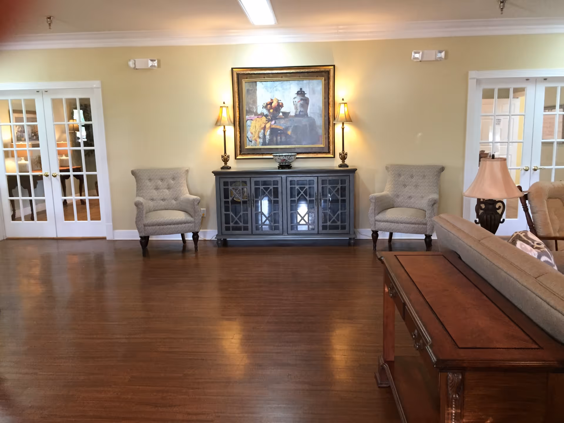 A cozy living room with wooden flooring, two upholstered armchairs flanking a dark wooden cabinet with glass doors, a framed painting above the cabinet, two wall lamps on either side of the painting, a beige sofa partially visible on the right, and a wooden side table with a lamp. French doors with glass panes are visible on both sides of the room.