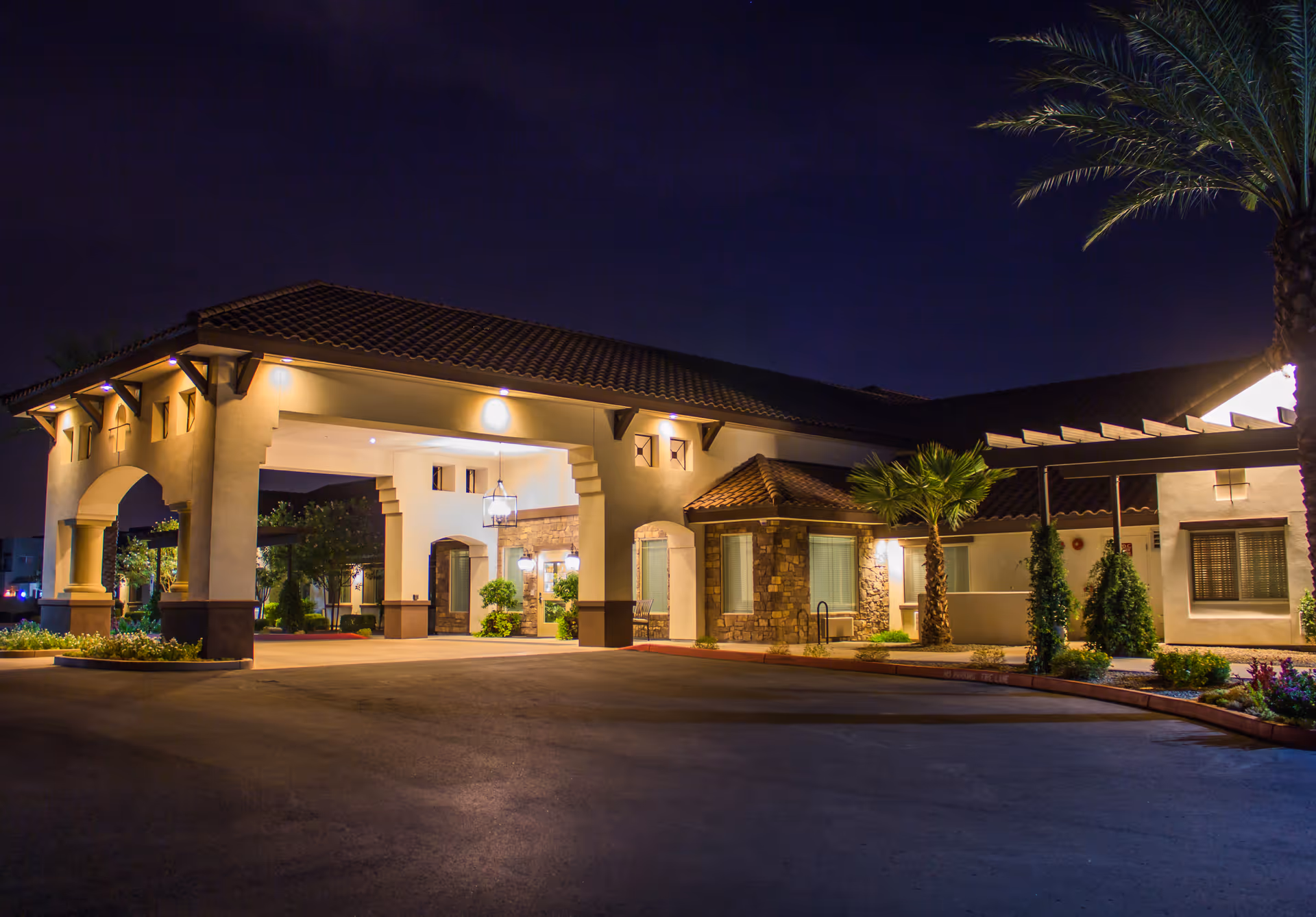 Night view of the entrance to a senior living facility with a covered driveway, exterior lights illuminating the building, palm trees, and landscaped greenery.