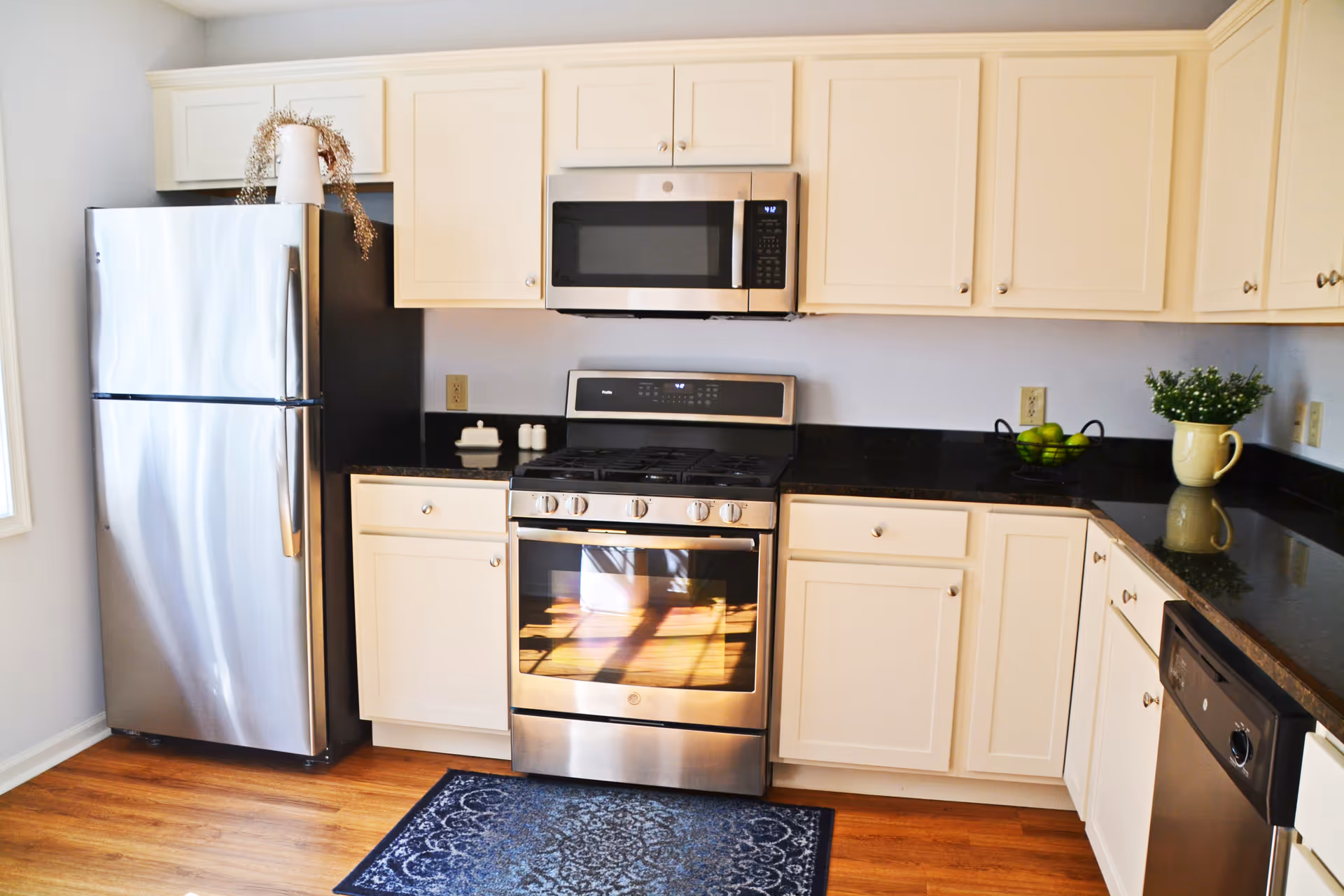 A modern kitchen with white cabinets, a stainless steel refrigerator, stove, microwave, and dishwasher. The countertop is black granite with a small green plant in a yellow pot and a bowl of green apples. There is a blue patterned rug on the wooden floor in front of the stove.
