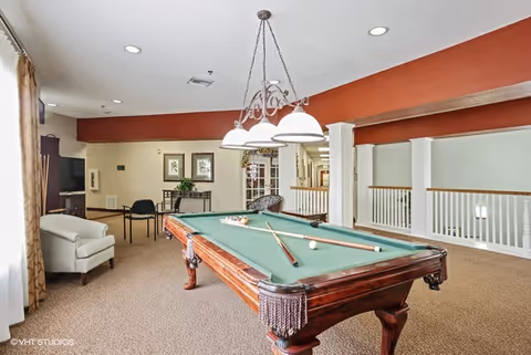 Communal game room featuring a pool table, overhead lighting, seating and a TV in a senior living facility.