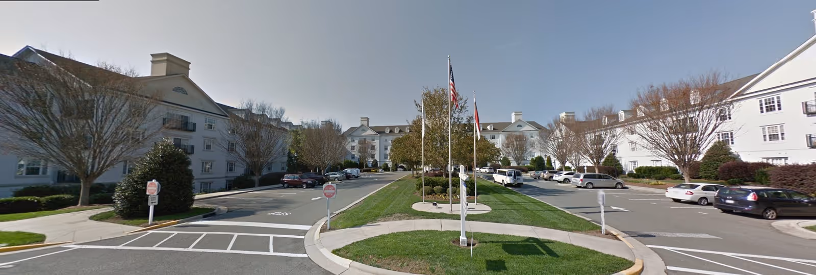 Front driveway and parking area of a large white senior living building with flagpoles and landscaped median.