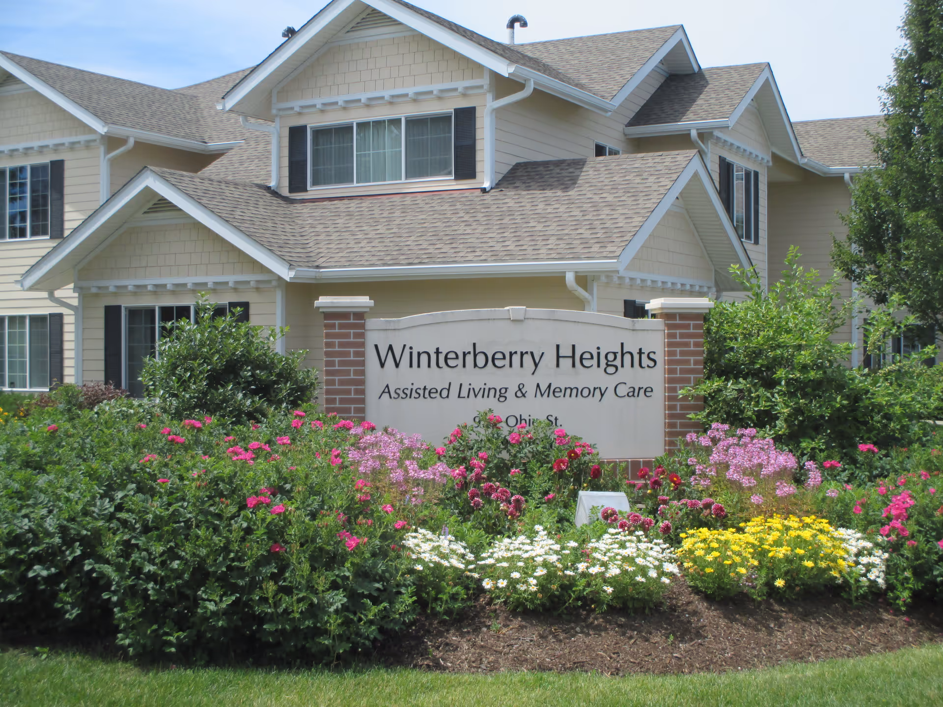 Exterior view of Winterberry Heights Senior Living facility with a large sign in front that reads 'Winterberry Heights Assisted Living & Memory Care' surrounded by colorful flowers and greenery, with the building visible in the background under a clear sky.