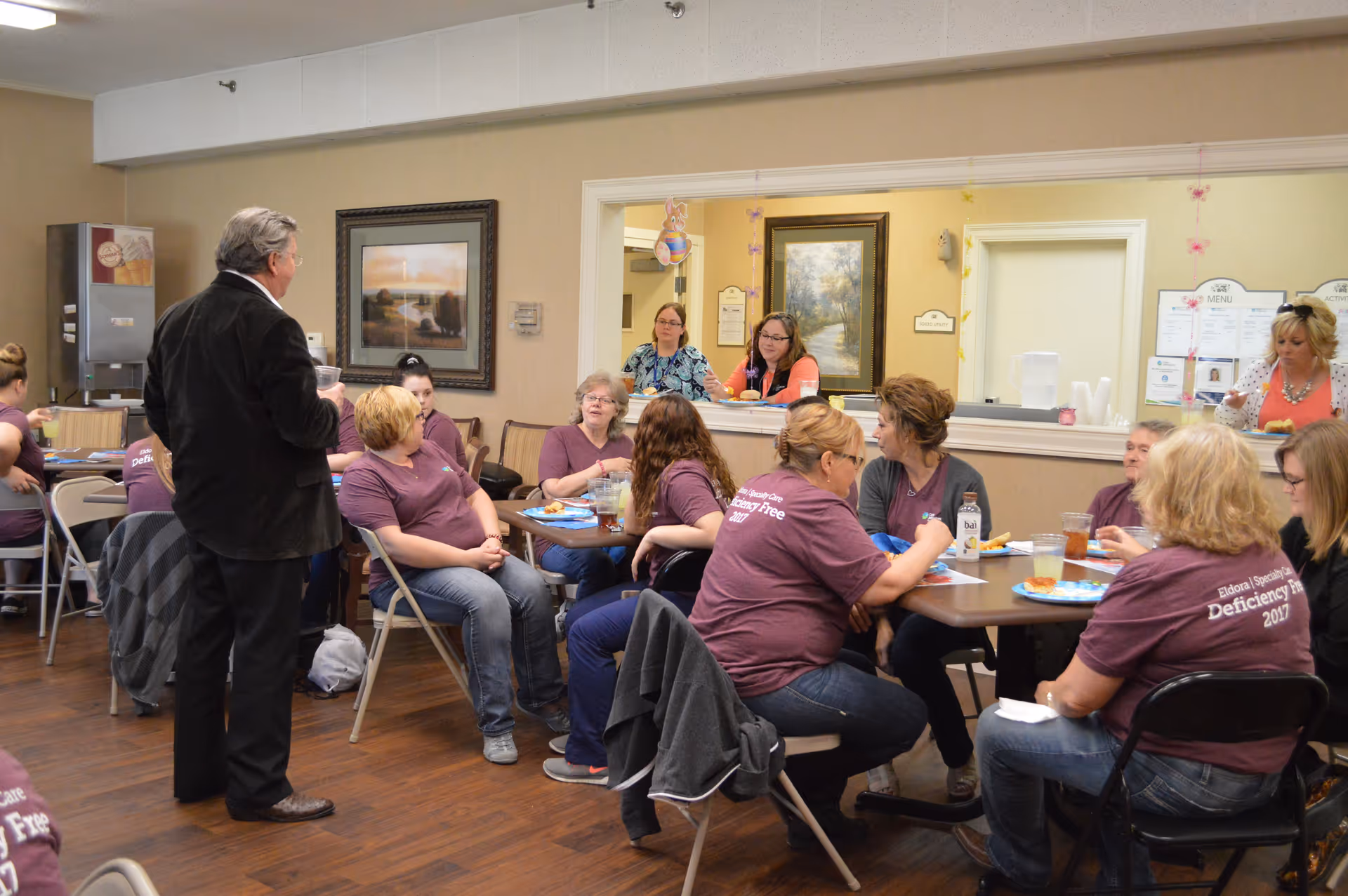 A group of people gathered in a common room with tables and chairs, some seated and eating while others stand and talk. The room has beige walls with framed pictures and a serving window where two women are standing behind the counter. Most of the seated people are wearing matching purple shirts with text on the back.