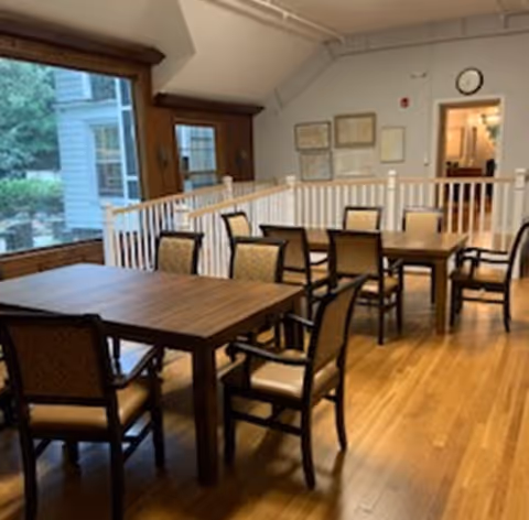 Interior view of a dining area with wooden floors, several wooden tables, and cushioned chairs. Large windows on the left side provide a view of greenery outside. The room has white walls with framed pictures and a clock above a doorway in the background.