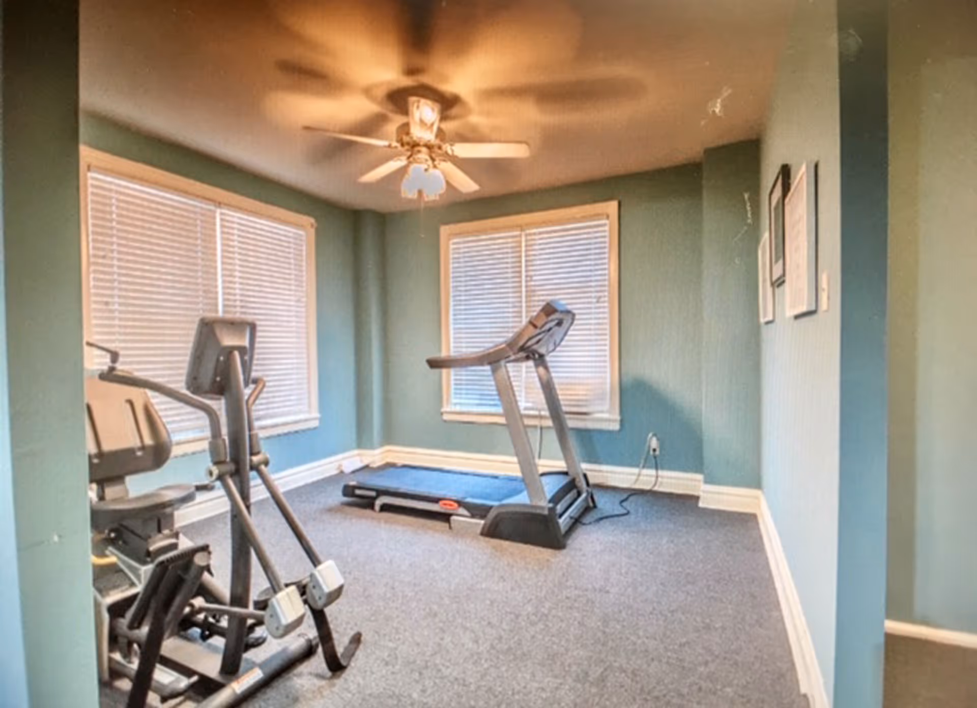 A small exercise room with teal walls and carpeted floor containing a treadmill and an elliptical machine. Two windows with closed blinds allow natural light into the room, and a ceiling fan with lights is mounted on the ceiling.