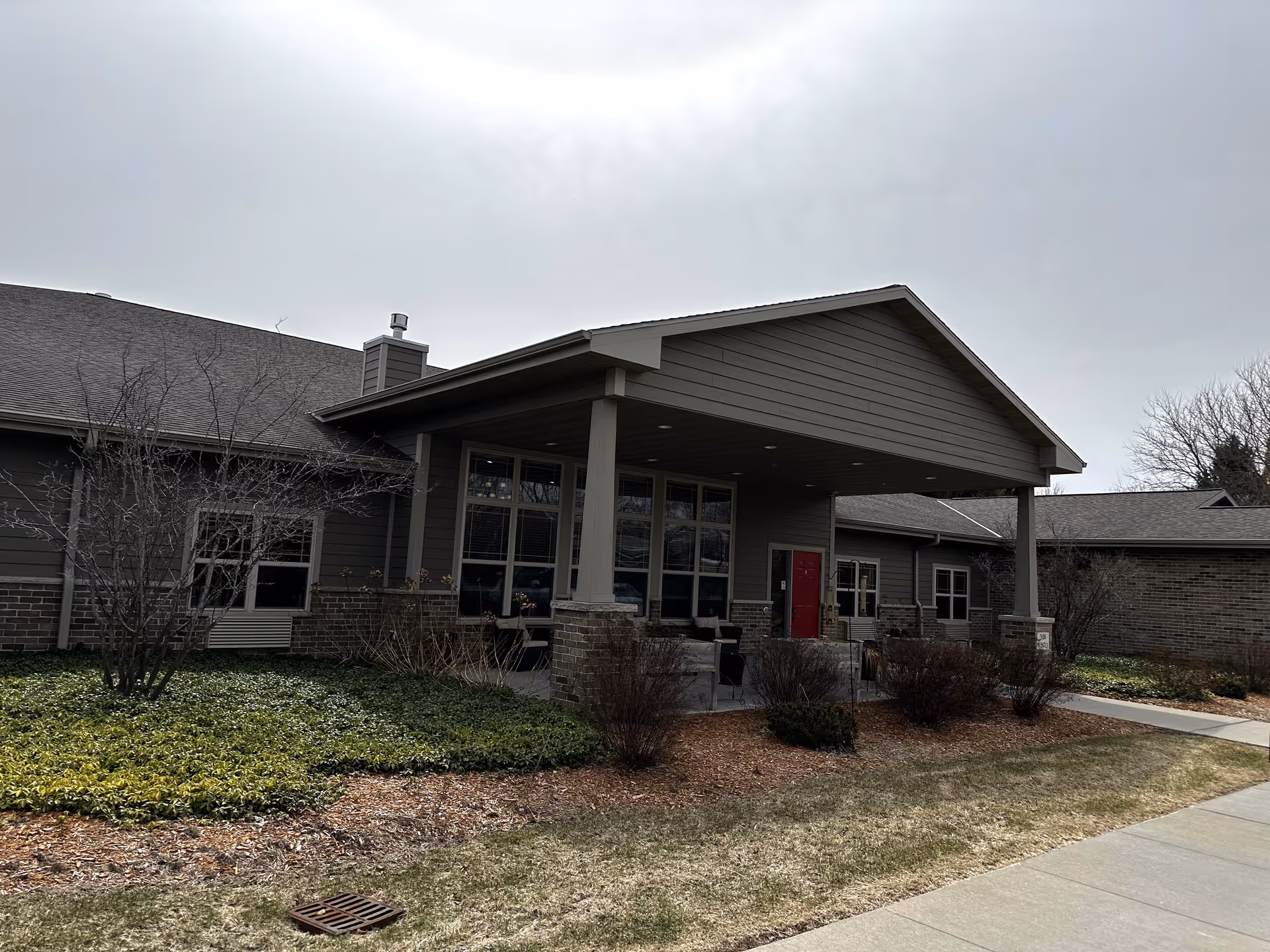 Exterior view of a single-story building with a covered entrance supported by columns. The building has gray siding with brick accents and several windows. There is a red door under the covered entrance. The surrounding area includes some bushes, grass, and a sidewalk leading to the entrance. The sky is overcast.