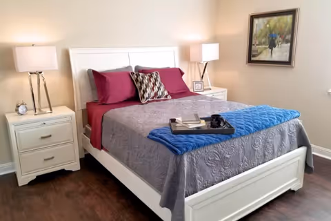 A neatly made bed with a white frame, gray quilt, blue throw blanket, and burgundy pillows in a bedroom. There are two white nightstands on either side of the bed, each with a lamp. A framed painting hangs on the beige wall to the right.