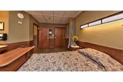 Private nursing facility bedroom with a made bed, wood furniture and headboard, bedside table with flowers, and a closed wooden door.