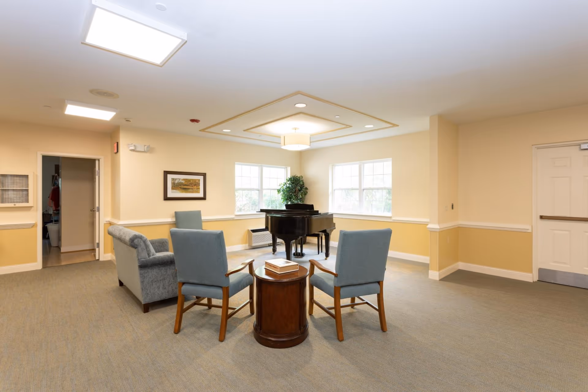 A bright and spacious common area with beige walls and carpeted floor featuring a black grand piano near two large windows. The room has a seating arrangement with two blue upholstered chairs and a small round wooden table between them, along with a blue upholstered loveseat. There is a framed picture on the wall and a potted plant near the piano. The ceiling has recessed lighting and a central light fixture.