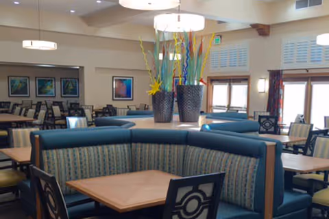 Interior view of a dining area with multiple tables and chairs. The foreground features a curved booth with blue and patterned upholstery surrounding a wooden table. In the center of the booth is a decorative arrangement of tall, colorful glass sculptures in two large vases. The background shows more tables and chairs, framed artwork on the walls, and large windows with curtains allowing natural light to enter.