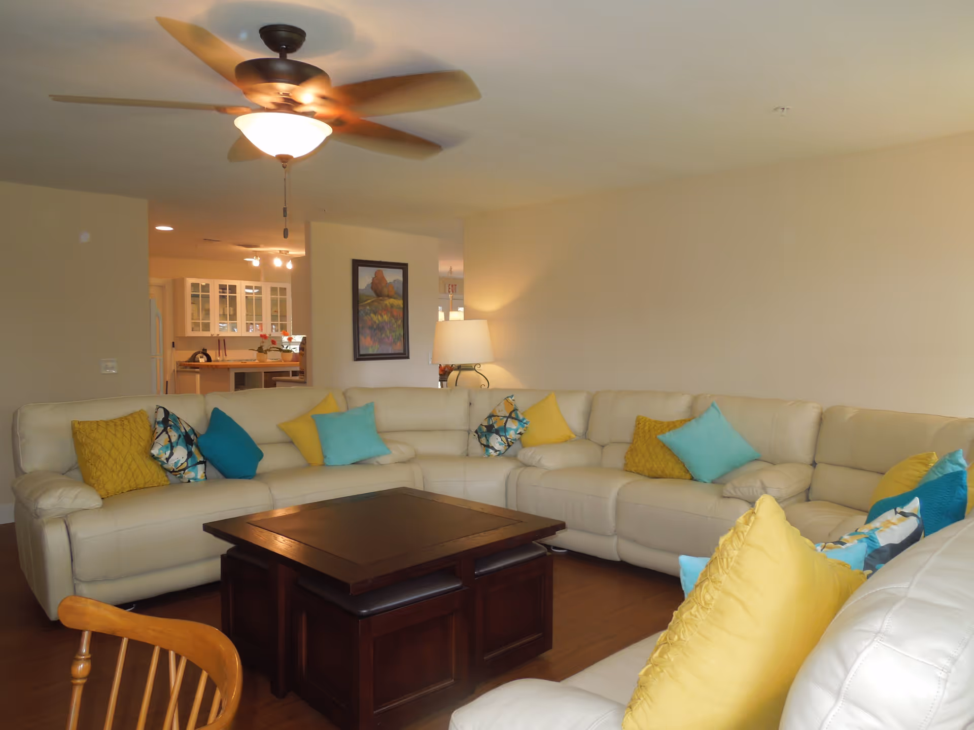A spacious living room with a large white sectional sofa adorned with yellow, turquoise, and patterned throw pillows. A dark wooden coffee table is centered in front of the sofa. The room has wooden flooring, a ceiling fan with a light, a table lamp, and a framed painting on the wall. The kitchen area is visible in the background.