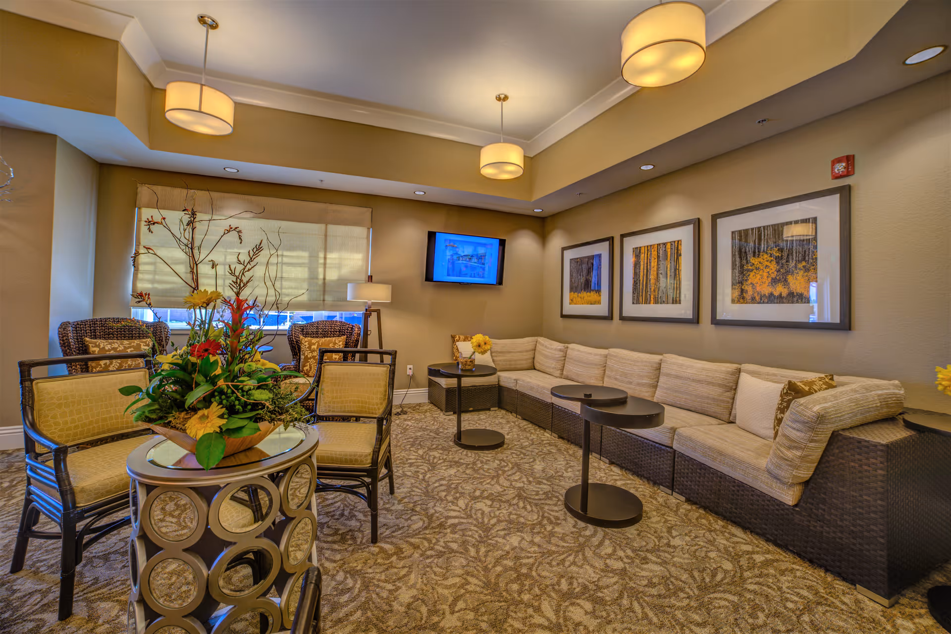 A cozy living room area with a long beige sectional sofa adorned with cushions, three framed nature-themed pictures on the wall, a flat-screen TV mounted on the wall, and a round table with a floral arrangement surrounded by four chairs. The room is warmly lit with ceiling lights and a floor lamp near the window with a beige shade.