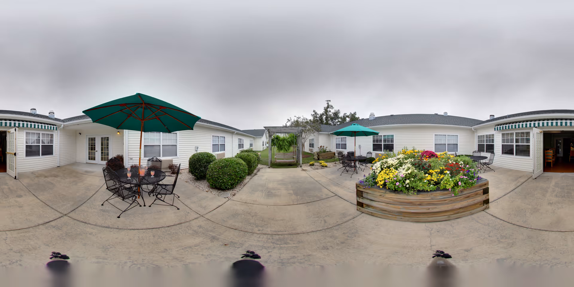 Outdoor courtyard area of a senior living facility with patio tables and chairs under green umbrellas, surrounded by well-maintained bushes and a large wooden planter filled with colorful flowers. The courtyard is enclosed by single-story white buildings with windows and doors, and the sky is overcast.