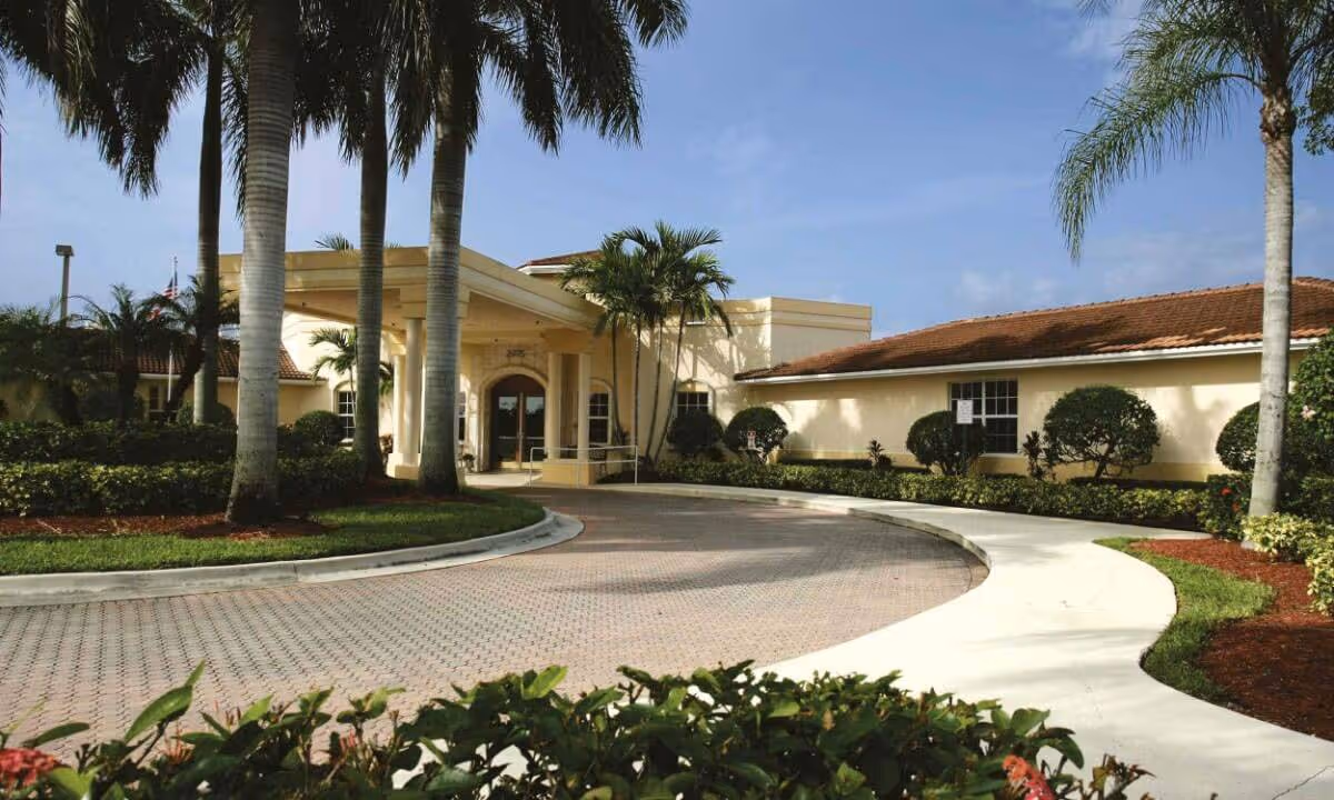 Entrance of a senior living building with a covered porte-cochere, circular driveway, palm trees, and landscaped grounds.