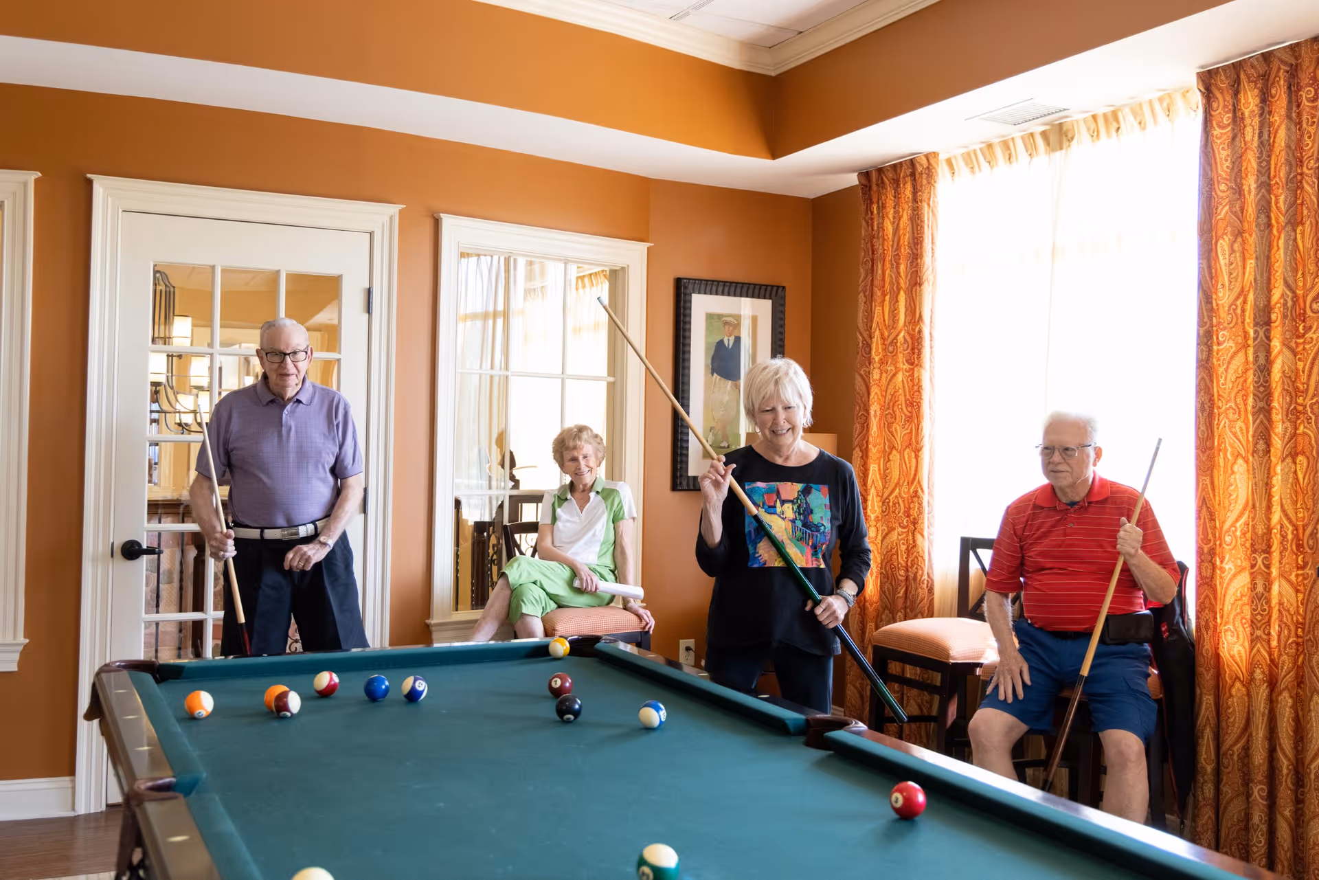 Four elderly people pose with pool cues around a billiards table in a bright recreation room.
