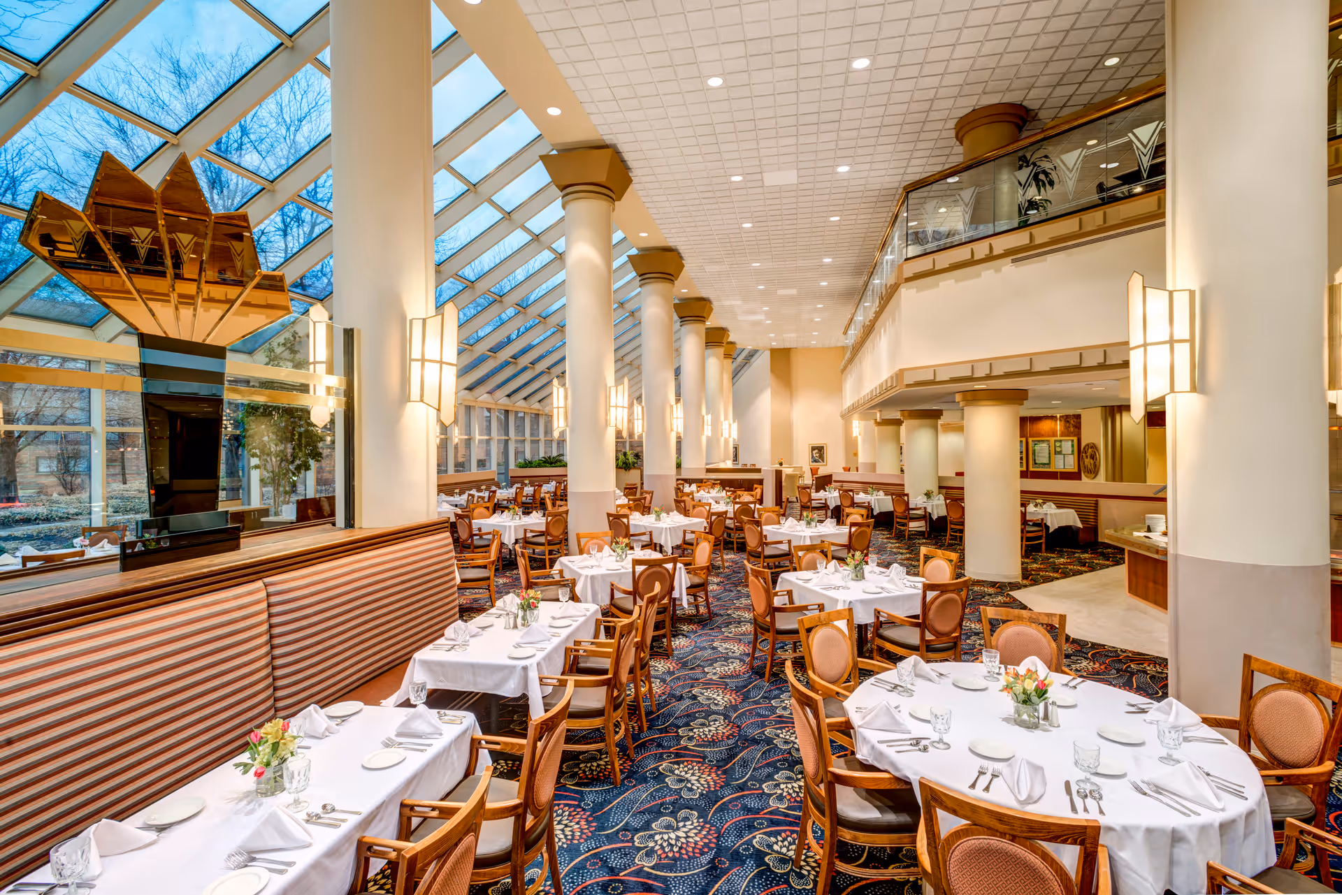 A spacious and elegant dining room with large windows and a glass ceiling allowing natural light to fill the space. The room features multiple round and rectangular tables covered with white tablecloths, set with glassware, plates, and silverware. Wooden chairs with cushioned seats surround the tables. There are tall white columns and warm wall sconces providing additional lighting. The carpet has a colorful floral pattern, and there is a decorative golden sculpture near the windows.