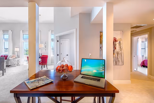 Open living area with a wooden desk holding a laptop, a glass bowl of apples, and a newspaper, with seating and doorways to rooms visible in the background.