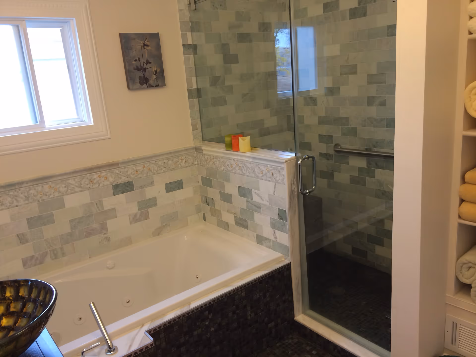 Bathroom with a white bathtub featuring jets, a glass-enclosed shower with tiled walls, a window, a small piece of wall art, and shelves with rolled towels.