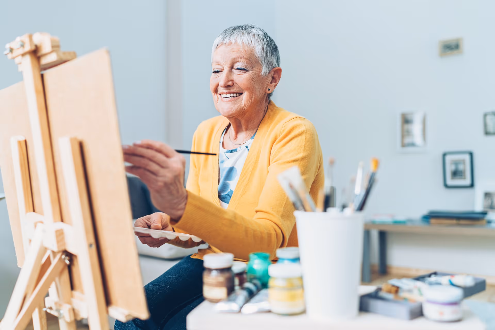 An elderly woman with short gray hair wearing a yellow cardigan is smiling while painting on a canvas in a bright room with art supplies and framed pictures on the wall.