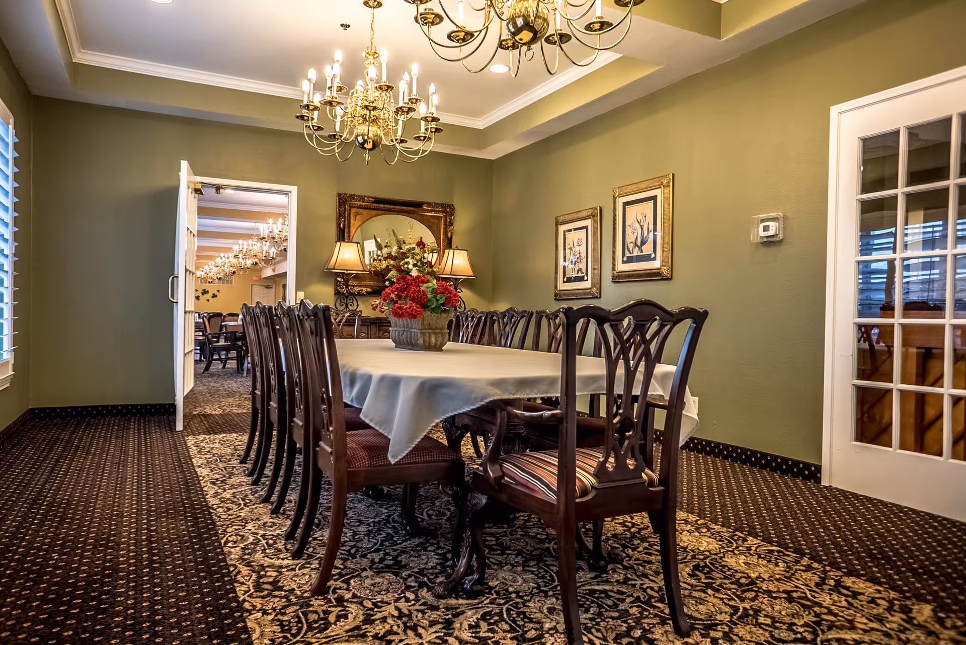 A formal dining room with a long table covered by a white tablecloth and surrounded by wooden chairs with patterned cushions. A floral centerpiece decorates the table. The room has green walls adorned with framed artwork and a large mirror above a sideboard with two lamps. A chandelier with multiple lights hangs from the ceiling. The carpet has an intricate pattern, and a door on the left opens to another room with more tables and chairs.