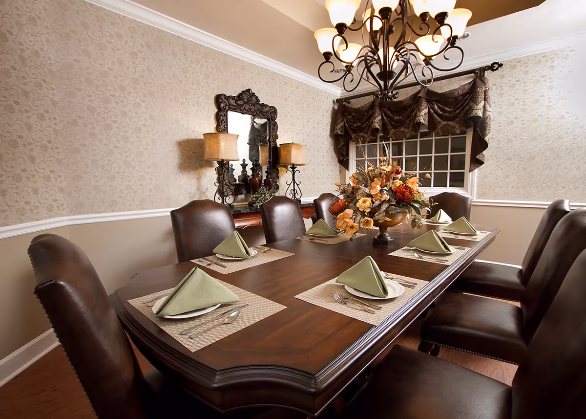 A formal dining room with a dark wooden table set for six people. Each place setting has a white plate, silverware, and a green folded napkin. The room features patterned wallpaper, a large decorative mirror, two table lamps with beige shades, a floral centerpiece on the table, and a chandelier overhead. A window with dark, ornate curtains is visible in the background.
