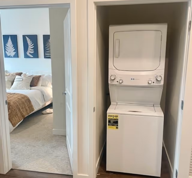 Stacked washer and dryer in a closet off a hallway with a bedroom visible through an open door.