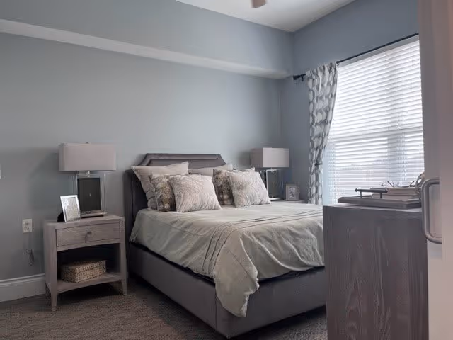 A neatly made bed with multiple pillows in a softly lit bedroom. The room features light gray walls, a window with patterned curtains, two bedside tables each with a lamp, and a small framed photo on one table. The floor is carpeted and there is a dresser partially visible on the right side.
