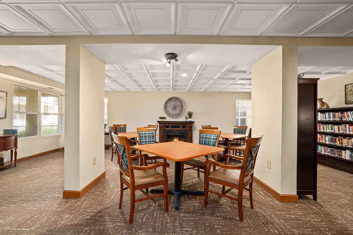 Communal dining area with wooden tables and upholstered chairs around a fireplace and bookshelves in a bright interior space.
