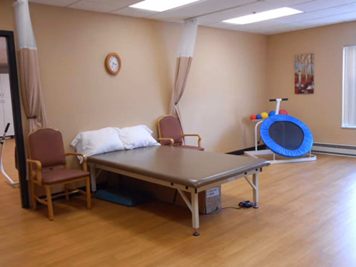 A therapy or exercise room with a padded treatment table, two wooden chairs with armrests, a small trampoline, and colorful exercise balls. The room has beige walls, a clock on the wall, a window with blinds, and a painting. The floor is wooden.